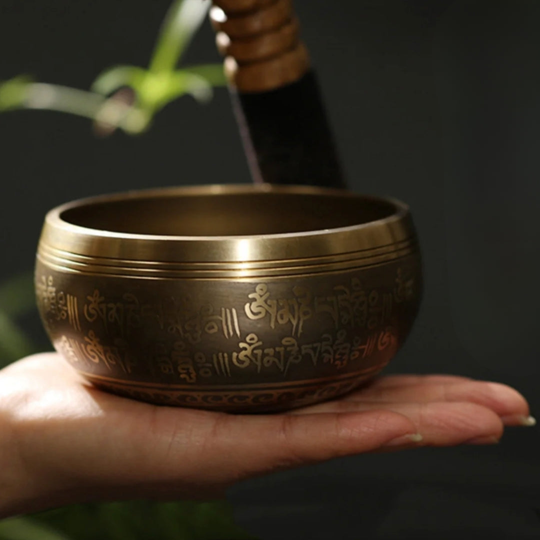 Image of a handcrafted CalmEcho Bowl Tibetan singing bowl, made in Nepal. The bowl is golden with an engraved traditional internal design, accompanied by a wooden striker with a padded end and a decorative blue and gold cushion. The set is used for meditation, yoga, and mindfulness practices, emitting relaxing and resonant sounds.