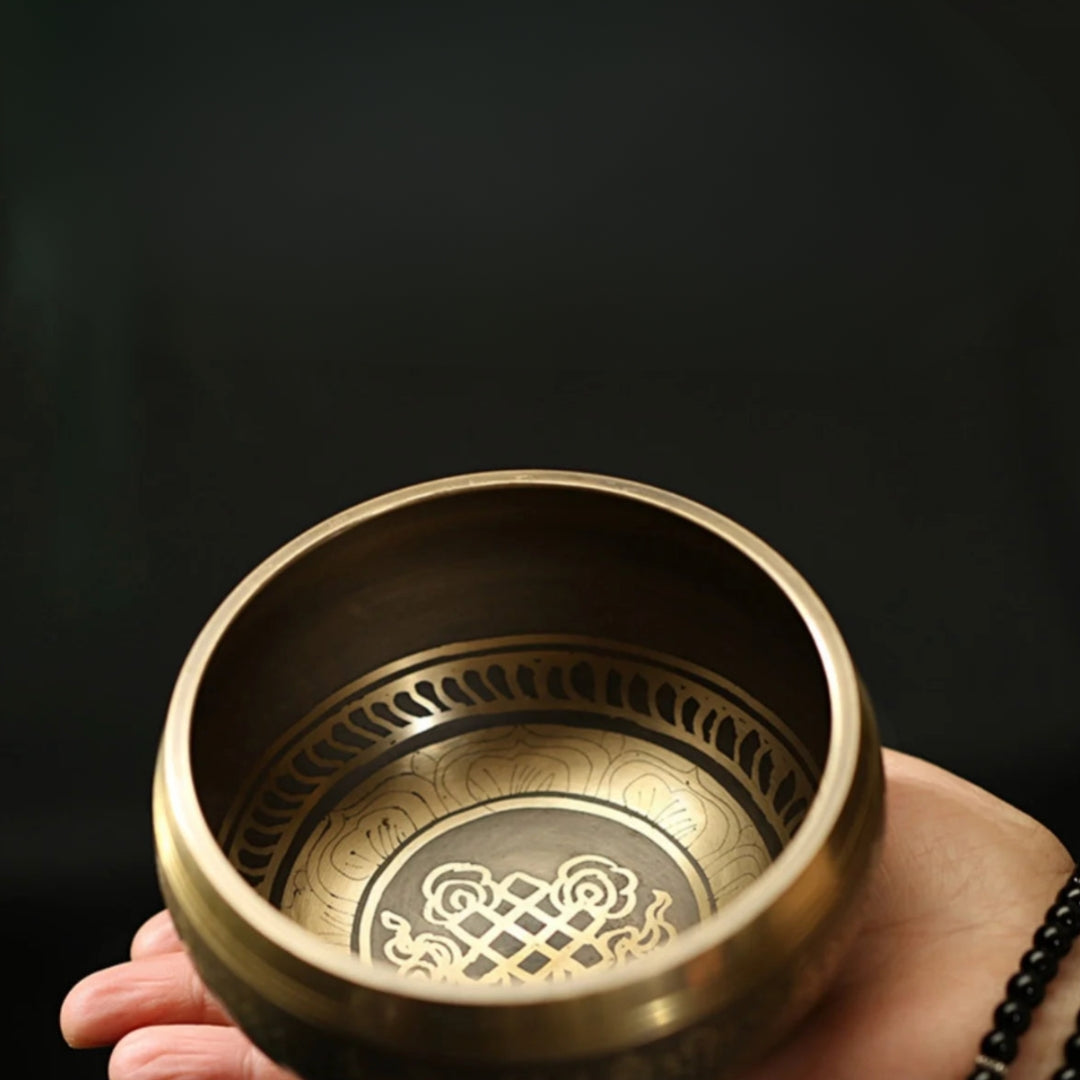 Image of a handcrafted CalmEcho Bowl Tibetan singing bowl, made in Nepal. The bowl is golden with an engraved traditional internal design, accompanied by a wooden striker with a padded end and a decorative blue and gold cushion. The set is used for meditation, yoga, and mindfulness practices, emitting relaxing and resonant sounds.