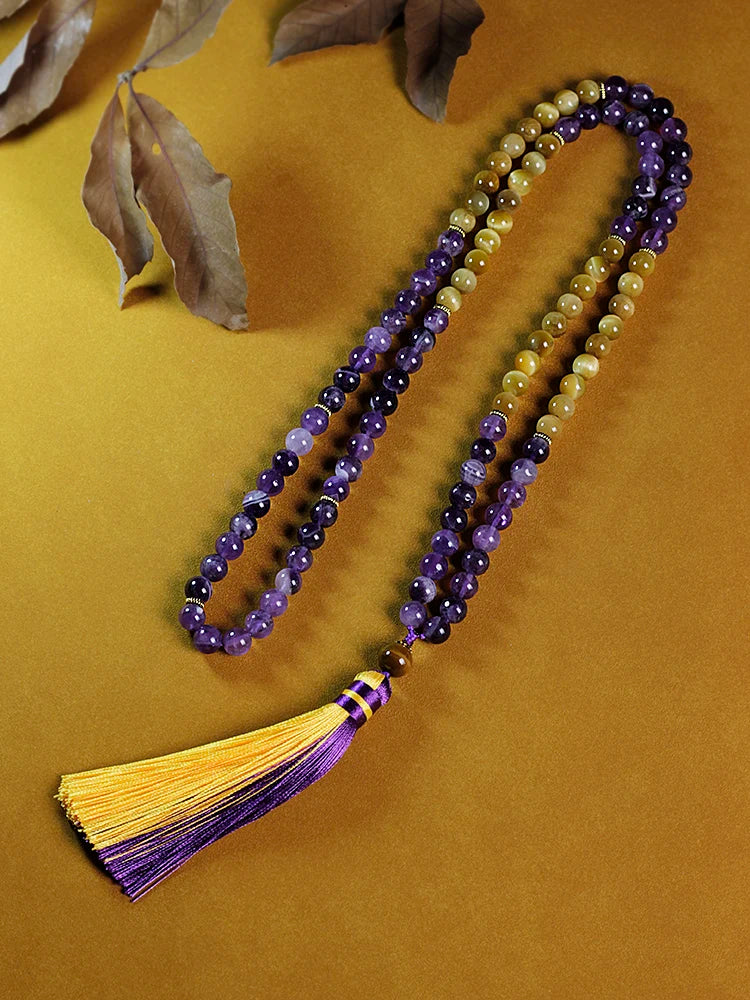 Image of the Ethereal Harmony Mala, a 108-bead meditation necklace made with purple amethyst and golden tiger eye stones. The mala features an elegant dual-color tassel in purple and yellow, coiled beautifully on a green leaf, highlighting its natural and spiritual essence. Ideal for meditation, yoga, or as a meaningful accessory.