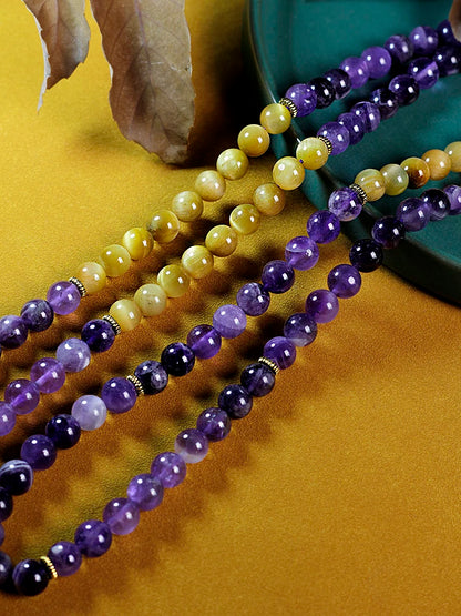 Image of the Ethereal Harmony Mala, a 108-bead meditation necklace made with purple amethyst and golden tiger eye stones. The mala features an elegant dual-color tassel in purple and yellow, coiled beautifully on a green leaf, highlighting its natural and spiritual essence. Ideal for meditation, yoga, or as a meaningful accessory.