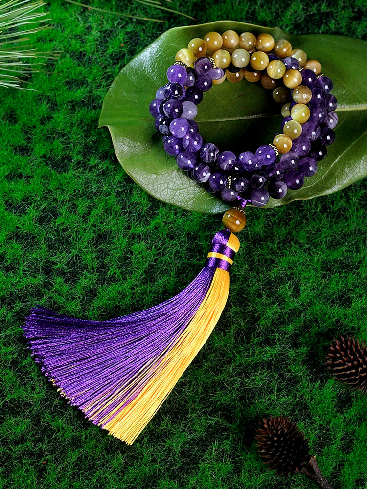 Image of the Ethereal Harmony Mala, a 108-bead meditation necklace made with purple amethyst and golden tiger eye stones. The mala features an elegant dual-color tassel in purple and yellow, coiled beautifully on a green leaf, highlighting its natural and spiritual essence. Ideal for meditation, yoga, or as a meaningful accessory.