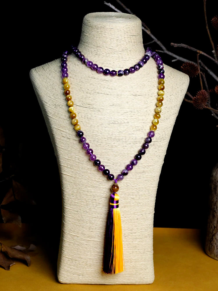 Image of the Ethereal Harmony Mala, a 108-bead meditation necklace made with purple amethyst and golden tiger eye stones. The mala features an elegant dual-color tassel in purple and yellow, coiled beautifully on a green leaf, highlighting its natural and spiritual essence. Ideal for meditation, yoga, or as a meaningful accessory.