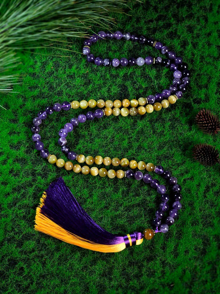 Image of the Ethereal Harmony Mala, a 108-bead meditation necklace made with purple amethyst and golden tiger eye stones. The mala features an elegant dual-color tassel in purple and yellow, coiled beautifully on a green leaf, highlighting its natural and spiritual essence. Ideal for meditation, yoga, or as a meaningful accessory.